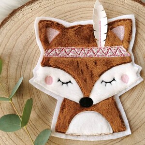Könnte beinhalten: Ein braunes und weißes Plüsch-Fuchs-Applik mit einem rosafarbenen Stirnband und einer weißen Feder. Der Fuchs hat geschlossene Augen und eine schwarze Nase.