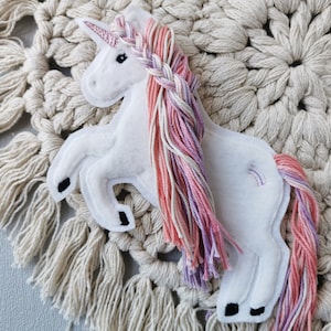 Könnte beinhalten: Ein weißes Filz-Einhorn mit einem rosa Glitzerhorn und einer geflochtenen Mähne aus rosa, pfirsichfarbenem und lavendelfarbenem Garn. Das Einhorn hat schwarze Hufe und ein kleines Regenbogen-Detail an der Seite. Das Einhorn wird auf einem cremefarbenen Makramee-Hintergrund präsentiert.