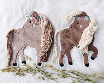 May include: Two brown plush horse appliques with embroidered floral details and yarn hair. One horse has a cream-colored mane and tail, while the other has a brown mane and tail.