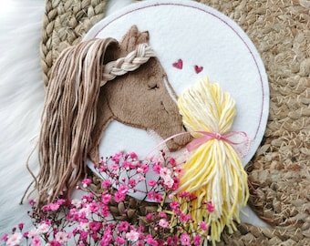 May include: Embroidered felt wall art featuring a brown horse with a braided mane and a yellow-blonde girl with a pink ribbon. The horse and girl are facing each other with two small pink hearts above them.