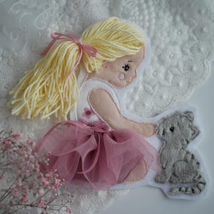Könnte beinhalten: Eine Filzpuppe mit gelben Garnhaaren und einem rosa Tüllrock, zusammen mit einer grauen Filzkatze. Die Puppe trägt ein weißes Kleid mit einer rosa Blume, vor einem weißen Spitzengrund.
