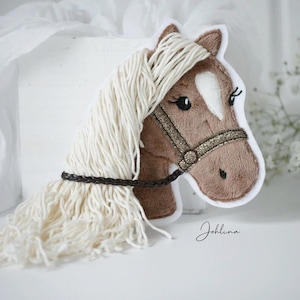 Könnte beinhalten: Ein Plüschpferdekopf mit cremefarbener Mähne und braunem Körper. Das Pferd hat einen goldglitzernden Halfter und einen dunkelbraunen geflochtenen Riemen. Das Bild enthält die handschriftliche Signatur "Johlina".