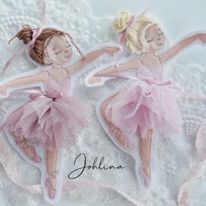 Könnte beinhalten: Zwei rosa Filz-Ballerina-Ornamente mit Tüllröcken und Schleifen. Eine hat braune Haare, die andere blonde. Beide befinden sich in einer Tanzpose, die Arme sind ausgestreckt. Das Wort "Johlina" ist kursiv geschrieben.