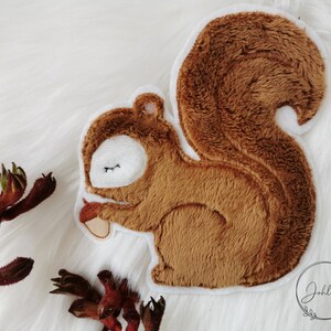 Könnte beinhalten: Ein braunes Plüsch-Eichhörnchen-Applik mit weißem Umriss. Das Eichhörnchen hält eine kleine braune Eichel in seiner Pfote. Das Applik ist auf weißem Hintergrund.