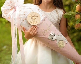 May include: A pink and white school cone with a cat applique and the name "Paulina" in gold glitter. The cone is decorated with gold polka dots, rainbows, and a wooden tag that says "Schulkind 2023".