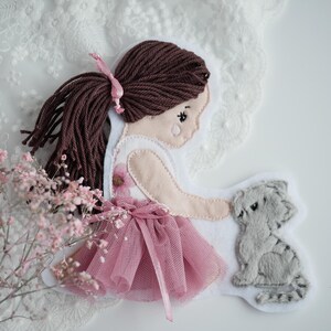 Könnte beinhalten: Handgefertigte Filzapplikation mit einem Mädchen in einem rosa Tutu-Kleid mit braunem Garnhaar und einem rosa Band. Das Mädchen interagiert mit einer grauen Filzkatze. Rosa Blumen und Spitze im Hintergrund.