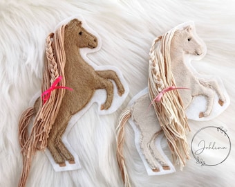 May include: Two felt horse appliques with braided manes and pink ribbons. The horses are brown and beige and are standing on their hind legs.