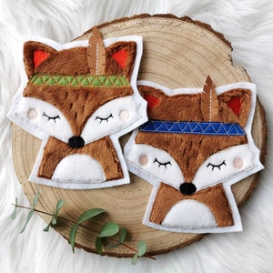 Könnte beinhalten: Zwei braune Plüsch-Fuchs-Applikationen mit weißen Gesichtern und geschlossenen Augen. Jeder Fuchs trägt ein Stirnband mit einer Feder und einem geometrischen Muster. Ein Stirnband ist grün und das andere ist blau.