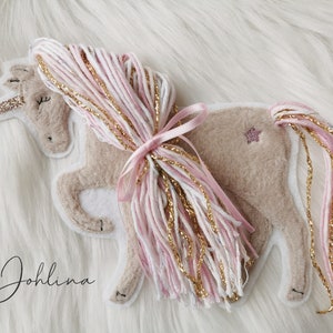 Könnte beinhalten: Ein beigefarbener Filz-Einhorn-Applikator mit einer rosa und goldenen Garnmähne und einem Schwanz. Das Einhorn hat ein glitzerndes goldenes Horn und einen rosa Stern an der Seite. Der Text "Johliina" ist in der linken unteren Ecke des Bildes sichtbar.