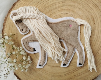 May include: A brown felt horse applique with a white yarn mane and tail. The horse is standing on a white background.