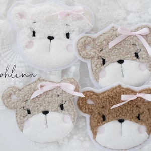 Könnte beinhalten: Vier Plüsch-Bärenkopf-Applikationen mit gestickten Gesichtern und rosa Schleifen. Die Bären sind in Weiß- und Brauntönen gehalten.