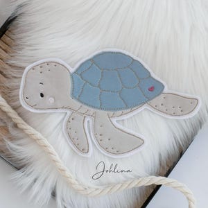 Könnte beinhalten: Eine blaue und beige Filzapplikation einer Schildkröte mit einem rosa Herzen auf ihrem Panzer. Die Schildkröte befindet sich auf einer weißen, flauschigen Oberfläche mit einem beigefarbenen Seil.
