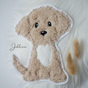 Könnte beinhalten: Ein flauschiger, hellbrauner Welpen-Applikation mit weißem Rand und einem herzförmigen weißen Fleck. Der Welpe hat schwarze Augen und Nase. Das Wort "Johlina" ist in Kursivschrift auf der linken Seite geschrieben.