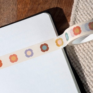 Peut inclure: Un rouleau de ruban adhésif washi présentant un motif répétitif de fleurs simples à cinq pétales dans des tons sourds d'orange, de violet, de marron, de bleu sarcelle et de jaune sur un fond crème, partiellement déroulé sur une surface blanche.