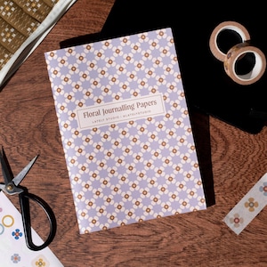 Könnte beinhalten: Ein rechteckiger Papierblock mit einem sich wiederholenden Muster aus weißen und braunen Blumen auf hellviolettem Hintergrund. Ein weißes Etikett in der Mitte zeigt den Text Floral Journalling Papers, LATELY STUDIO und @LATELYSTUDIO in Dunkelbraun.