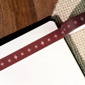 May include: A roll of dark maroon washi tape featuring a repeating pattern of light pink four-pointed stars and simple five-pointed stars. The tape is partially unrolled and applied across the top edge of a blank white rectangular paper surface.