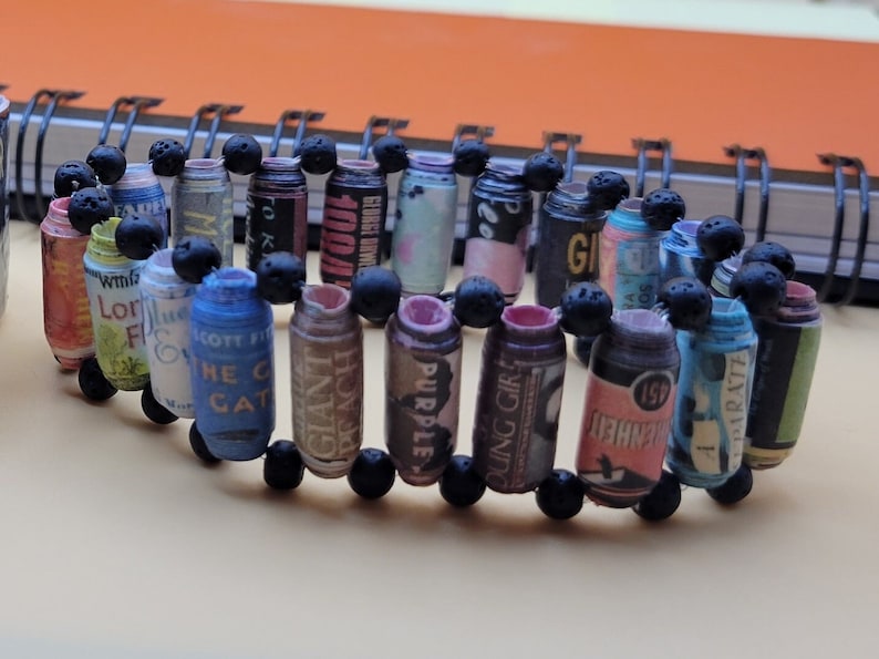 May include: A bracelet made of rolled up paper with different text and designs. The paper is rolled into small cylinders and strung together with black beads.