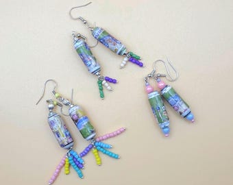 May include: Handmade earrings with cylindrical beads displaying a landscape design, complemented by colourful beads in pink, blue, yellow, and green. The earrings have silver-tone ear wires, presented on a light beige surface.