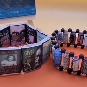 May include: Two book-themed bracelets. One bracelet is made with small book covers and the other is made with rolled-up pages of books and black beads.