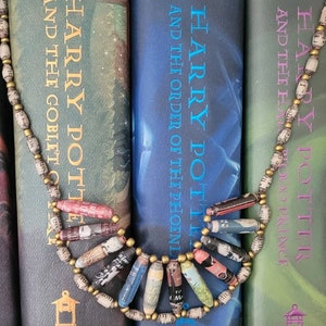 May include: Three Harry Potter books, volumes 4, 5, and 6, with a paper bead necklace in front of them. The books are green, blue, and green, and the necklace is made of brown and white paper beads.