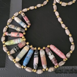 May include: A necklace made of rolled paper beads with gold accents. The beads are decorated with magazine clippings and text. The necklace is on a dark gray surface.