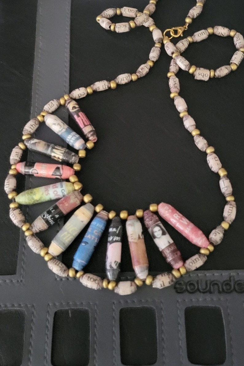 May include: A necklace made of rolled paper beads with gold accents. The beads are decorated with magazine clippings and text. The necklace is on a dark gray surface.