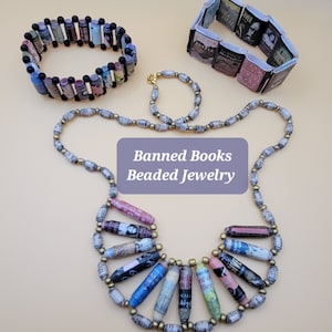 May include: A set of three necklaces made from rolled up pages of banned books. The necklaces are in different lengths and styles. The necklaces are made with gold and black beads. The necklaces are labeled "Banned Books Beaded Jewelry".