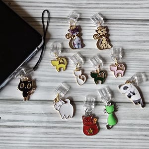 May include: A collection of cat-themed phone charms in various colors, including black, white, gold, green, and pink. Each charm features a different cat design and is attached to a clear plastic plug. The charms are displayed on a light-colored wooden surface.