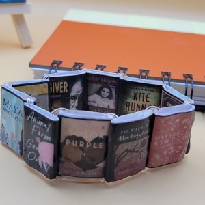 May include: A bracelet made of miniature book covers. The covers feature titles such as "The Giver", "Animal Farm", "The Diary of Anne Frank", "Kite Runner", "To Kill a Mockingbird", and "The Handmaid's Tale".