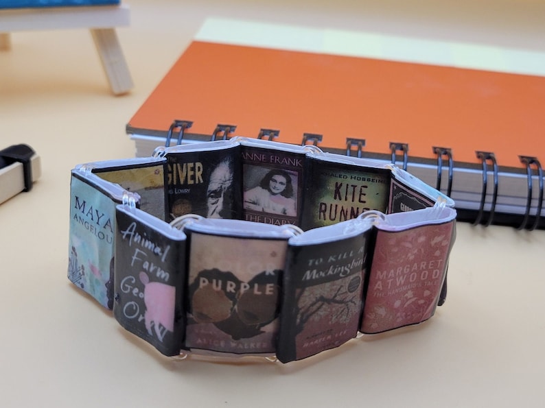 May include: A bracelet made of miniature book covers. The covers feature titles such as "The Giver", "Animal Farm", "The Diary of Anne Frank", "Kite Runner", "To Kill a Mockingbird", and "The Handmaid's Tale".