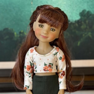 May include: A doll with long auburn hair and a floral print top. The doll has a fair complexion, rosy cheeks, and green eyes. She is wearing a white long-sleeved shirt with orange and pink floral patterns and a dark green skirt.