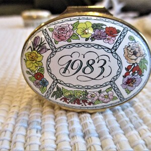 May include: Oval trinket box with a gold-tone rim and a floral design. The lid features a white background with colorful flowers and the year "1983" in the center. The box is set on a textured white surface.