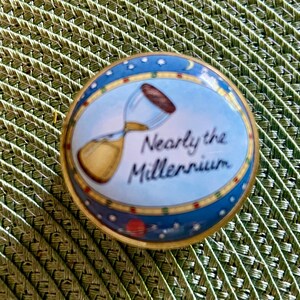 May include: A round, gold-rimmed trinket box with a light blue lid featuring a graphic of an hourglass and the text Nearly the Millennium. The border of the lid is decorated with small stars, a crescent moon, and a red planet against a dark blue sky.