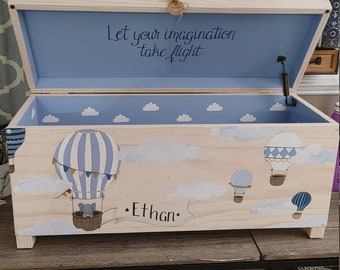 May include: A wooden toy chest with a blue and white hot air balloon design. The lid is open and the inside is painted blue with white clouds. The chest is decorated with the text "Let your imagination take flight" and "Ethan".