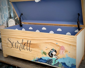 May include: A wooden toy chest with a blue interior and a mermaid design painted on the front. The chest is decorated with white clouds and the name "Scarlett" is written on the front.