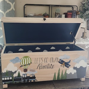 May include: A wooden toy chest with a blue interior and a painted design of a hot air balloon, airplane, and train. The chest is decorated with clouds and the words "Let's go on an Adventure".