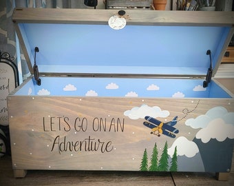 May include: A wooden toy chest with a light blue interior and a painted scene of a plane flying over mountains and trees. The chest has the words "Let's go on an adventure" painted on the front.