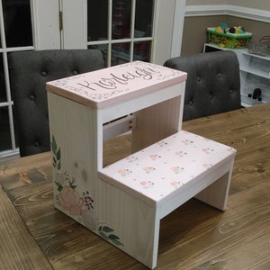 May include: A white wooden two-step stool with a pink top step. The top step has a floral design and the name "Karleigh" written in cursive. The bottom step has a floral pattern.
