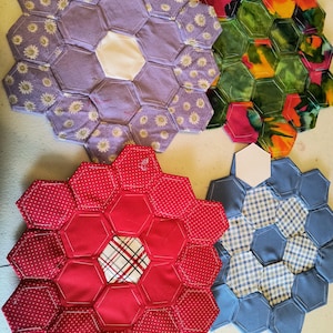 May include: Four handmade fabric coasters in a flower shape. One is purple with daisy print, another is red with white polka dots, a third is blue with a gingham pattern, and the last is green with a floral pattern. Each coaster is made of hexagonal fabric pieces.