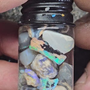 Puede incluir: Un pequeño frasco de vidrio lleno de varios trozos de ópalo. El ópalo es iridiscente y muestra una variedad de colores, incluyendo azul, verde y morado.