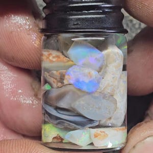 Puede incluir: Un pequeño frasco de vidrio lleno de piedras preciosas de ópalo iridiscente. El frasco tiene una tapa de rosca negra.