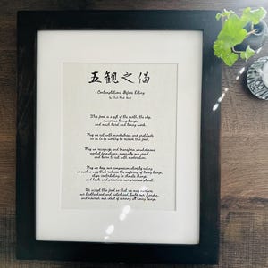 May include: A framed print with a black frame and a white mat. The print features a poem in black ink titled "Contemplations Before Eating" with the text "五觀之偈" in Chinese characters at the top. The poem is written in a cursive script and includes the following text: "This food is a gift of the earth, the sky, numerous living beings, and much hard and loving work. May we sit with mindfulness and gratitude so as to be worthy to receive this food. May we recognize and transform our greed, mental formations, especially our desires, and here to sit with moderation. May we bear our compassion for all beings in such a way that reduces the suffering of living beings, stops contributing to climate change, and helps preserve our precious planet. We accept this food as that we may nurture our brotherhood and sisterhood, build our strength, and nourish our diet of serving all living beings."