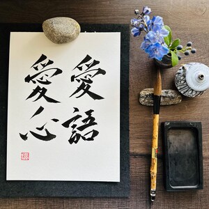 May include: A black and white calligraphy piece on white paper with the Chinese characters for "love" and "forever" written in black ink. The paper is mounted on a black mat and there is a small red seal in the lower left corner. The image also includes a small rock, a blue flower, a brush, and a black ink stone.