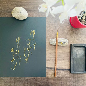 May include: A black sheet of paper with gold calligraphy on it. There is a small rock, a black ink stone, and a brush on a wooden surface.