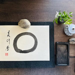 May include: A black ink circle drawn on white paper with the Japanese characters "夏休" written in black ink on the left side of the circle. The paper is on a black mat and there are various other items on a wooden surface, including a small potted plant, a stone, a black ink stone, and a brush.