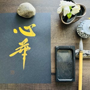 May include: A black calligraphy paper with gold ink calligraphy of the Japanese characters "春" and "心". The characters are written in a traditional style. There are other items in the image, including a stone, a small vase with white flowers, a small inkwell, and a brush.