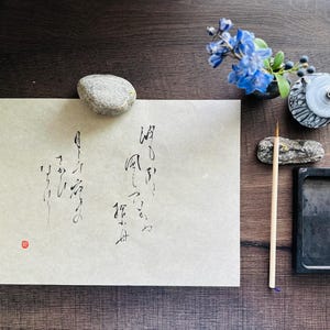 May include: A white sheet of paper with black calligraphy on it. The paper is on a wooden surface with a small rock, a blue flower, a small inkwell, and a brush.