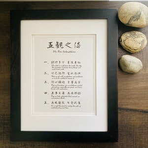 May include: A framed print with black and white text in both English and Japanese. The text reads "The Five Contemplations" and includes five verses in Japanese. The print is framed in a black frame and is sitting on a wooden surface.