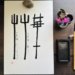 May include: Ink painting on white paper featuring three abstract black shapes and Japanese calligraphy. A small red stamp is in the lower left corner. A brush, ink stone, and small floral arrangement are on a wooden surface.