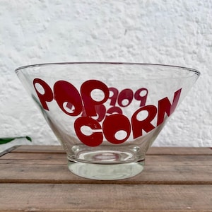 Op de afbeelding: Een heldere glazen kom met een brede, uitlopende rand en een ronde voet. De buitenkant van het glas is voorzien van het woord POPCORN in dikke, rode, afgeronde hoofdletters die in een gestileerd, modern lettertype rond de kom zijn geplaatst.